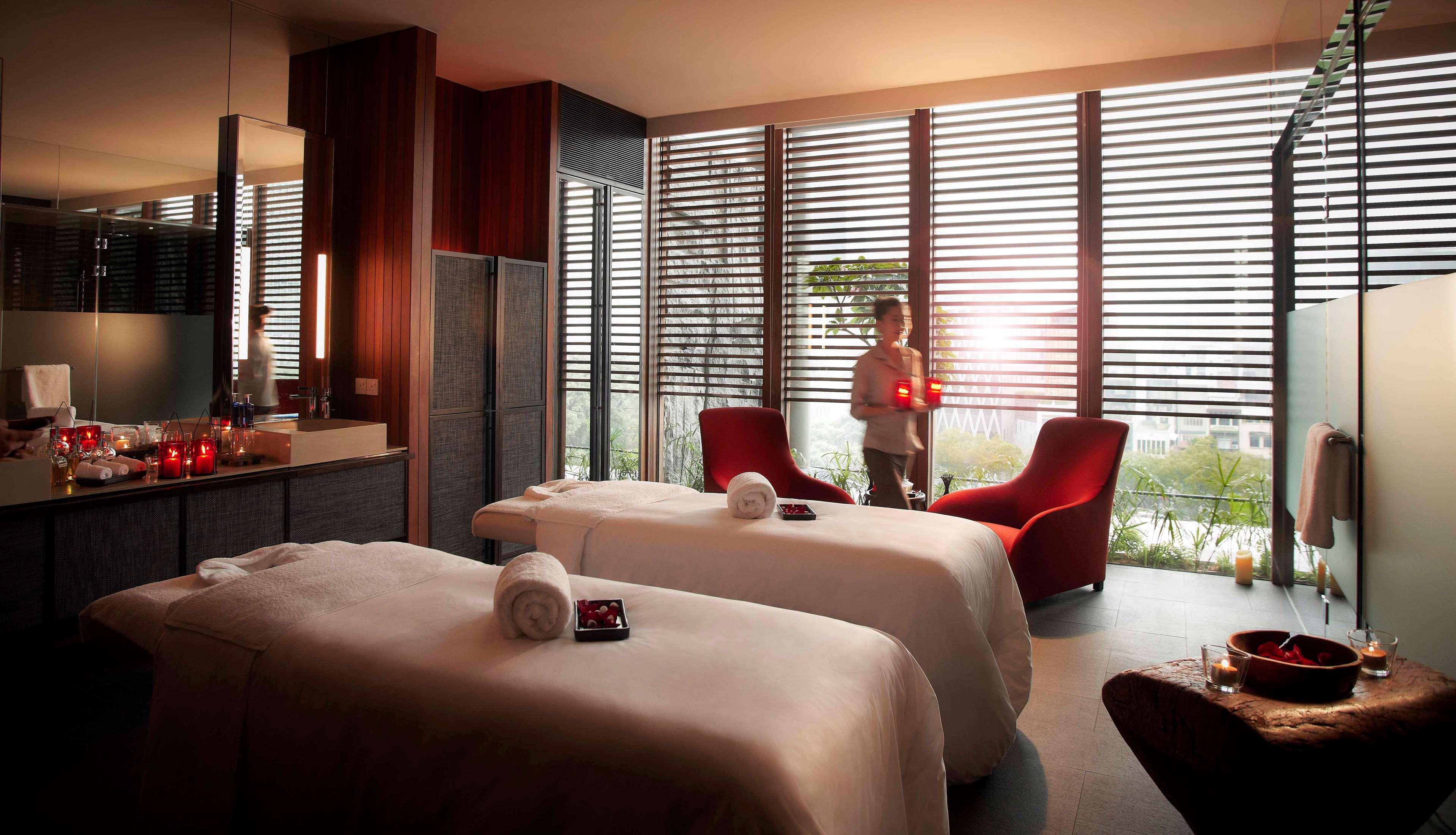 Parkroyal Collection Pickering, Сингапур Екстериор снимка The photo shows a tranquil spa room with a calming ambiance. There are two massage tables with white linens and neatly rolled towels placed on them. In the background, large windows with horizontal slats allow soft light to filter in, creating a sere