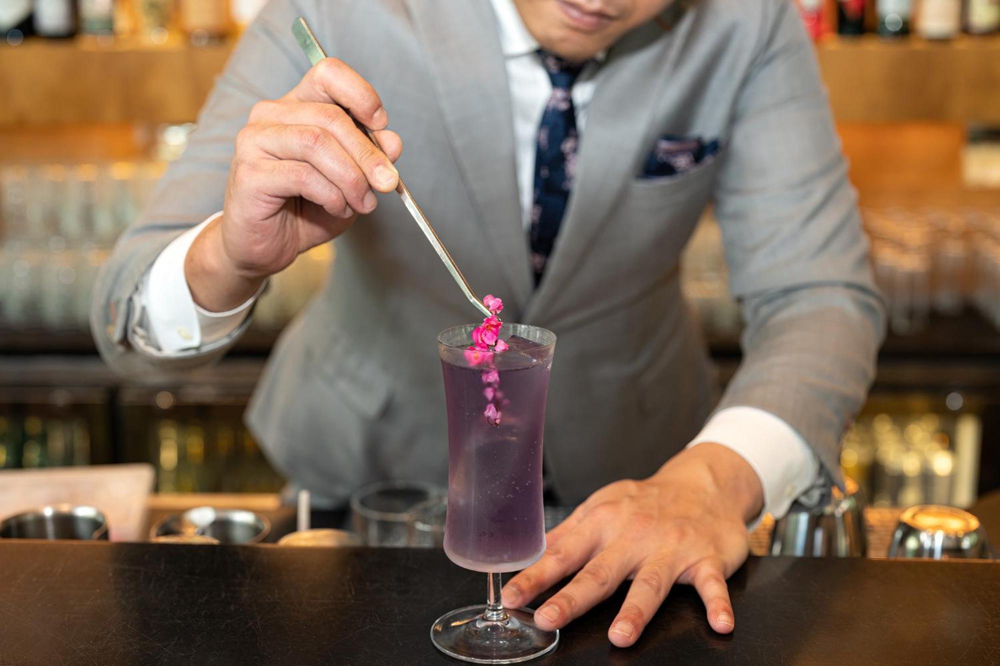 Parkroyal Collection Pickering, Сингапур Екстериор снимка The photo shows a bartender in a stylish gray suit preparing a cocktail. He is holding a long, thin tool, possibly a stirrer or a tweezer, and is carefully placing a small pink flower garnish into a tall glass filled with a vibrant purple drink. The 