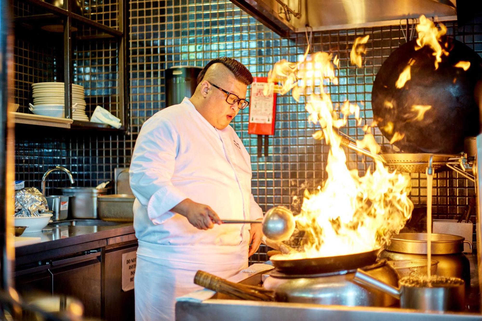 Parkroyal Collection Pickering, Сингапур Екстериор снимка The photo shows a chef in a kitchen, wearing a white chef's jacket and glasses. He is actively cooking in front of a large wok that is producing a dramatic surge of flames. The kitchen has a modern look, with tiled walls and a variety of kitchen equi