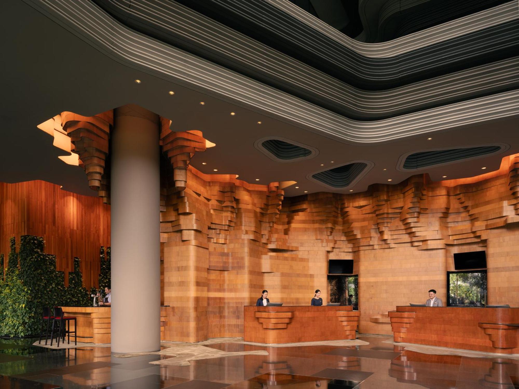 Parkroyal Collection Pickering, Сингапур Екстериор снимка The image shows a modern hotel or building lobby with a sophisticated design. The interior features richly textured wooden walls that resemble natural formations, creating a warm and inviting atmosphere. The reception area has several service counter