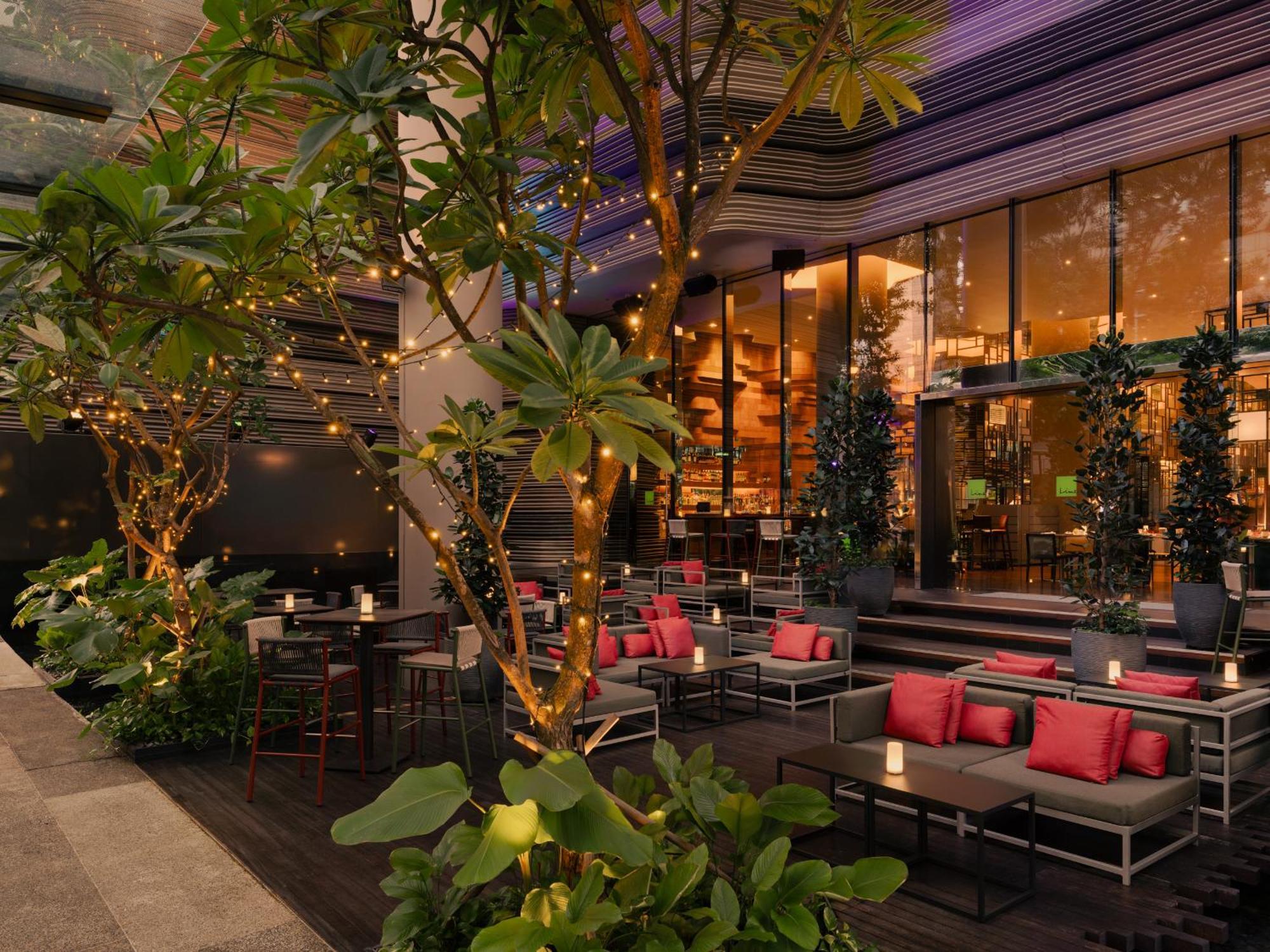 Parkroyal Collection Pickering, Сингапур Екстериор снимка The photo shows a stylish outdoor seating area of a restaurant or lounge. There are several tables and chairs arranged comfortably, featuring cushions in a vibrant pink color. Surrounding the seating area are lush green plants and trees, which add a 